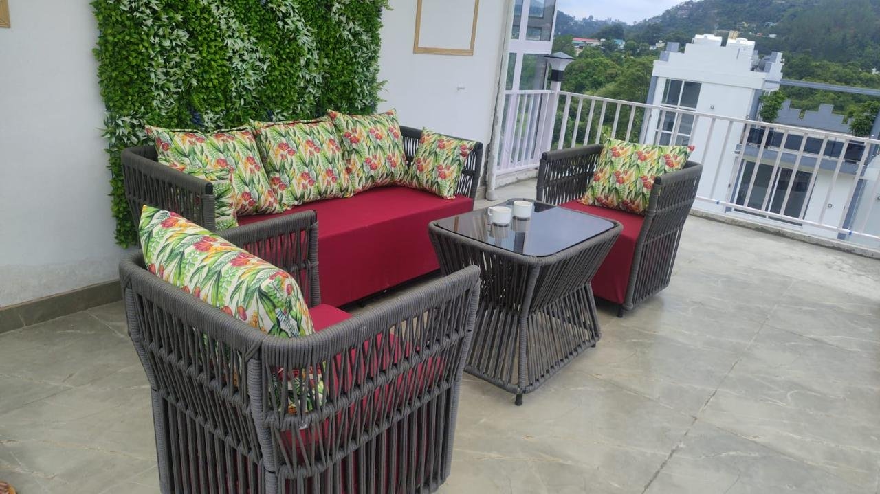 Hillside Social rooftop lounge with grey woven sofa, burgundy cushions, floral pillows, and green hill views